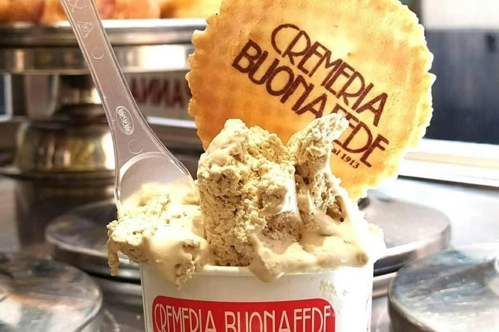 一份丰盛的奶油冰淇淋盛放在印有 Crèmeria Buona Fede 标志的白色罐子中。一块印有冰淇淋店名称的金黄色华夫饼盖在冰淇淋上，一把塑料勺子插在奶油中。